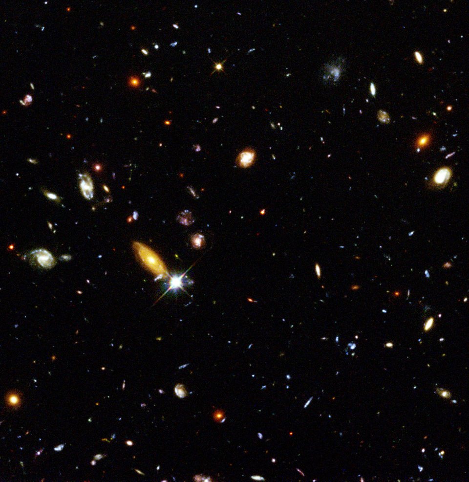 Hundreds of small galaxies of different shapes and colours, spirals, ellipses and specks of light scattered across a black field.