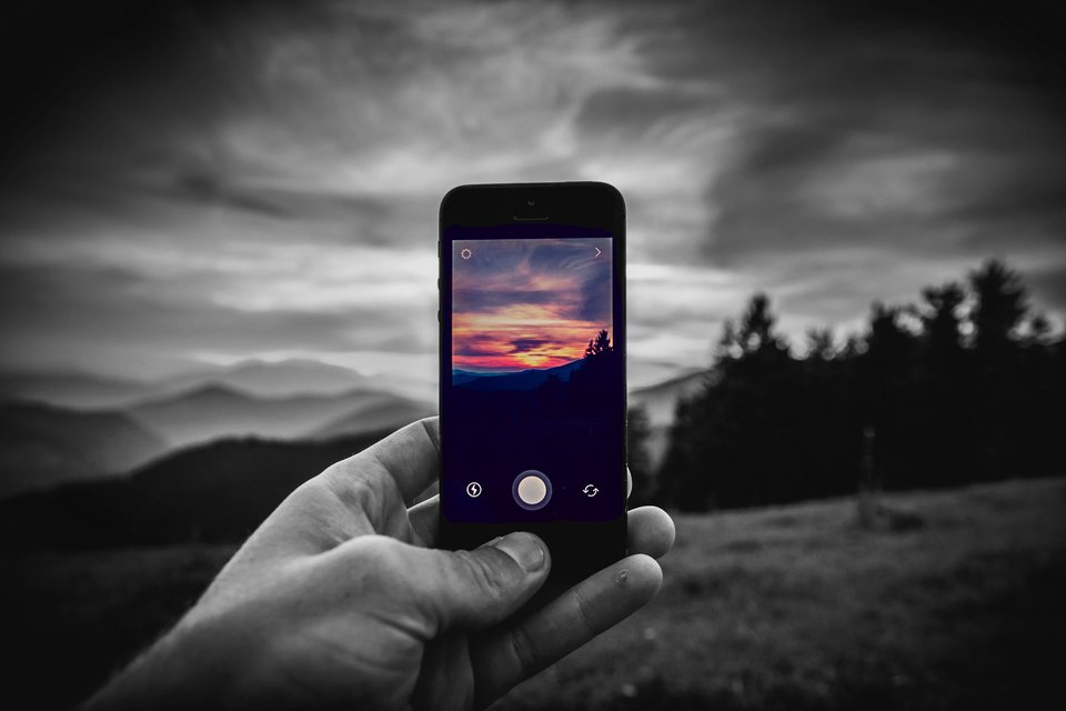 A hand holds up a smartphone showing a vivid orange sunset photo against the same view in black and white: hills and a dark forest edge.