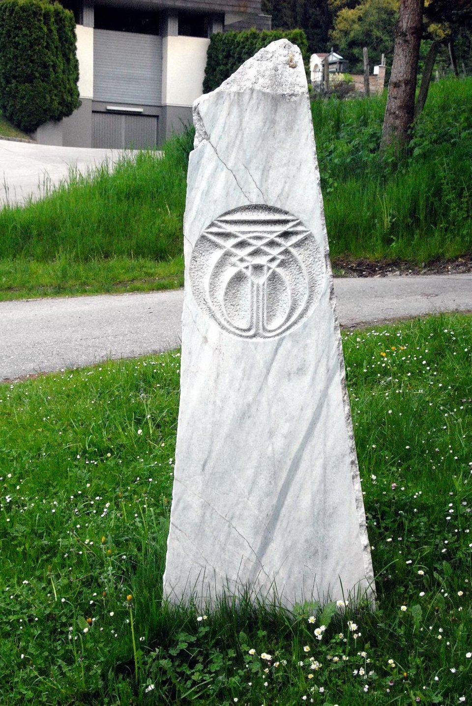 An upright slab of white marble beside a path and a grassy slope, carved with a round relief sign.